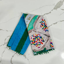 Load image into Gallery viewer, Decorative tea towel with colorful sprinkle cookies on blue and green striped background design on marble surface.