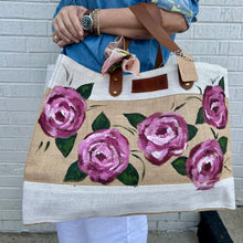 Load image into Gallery viewer, Person holding a tote bag with hand painted pink rose floral design against a white brick wall.