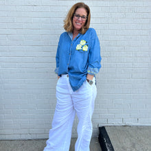 Load image into Gallery viewer, Woman wearing a blue denim shirt with floral painted flowers and white pants standing against a white brick wall.