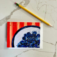 Greeting card of painting of blueberries on a white plate with a red background, next to a yellow pencil on a textured surface. 